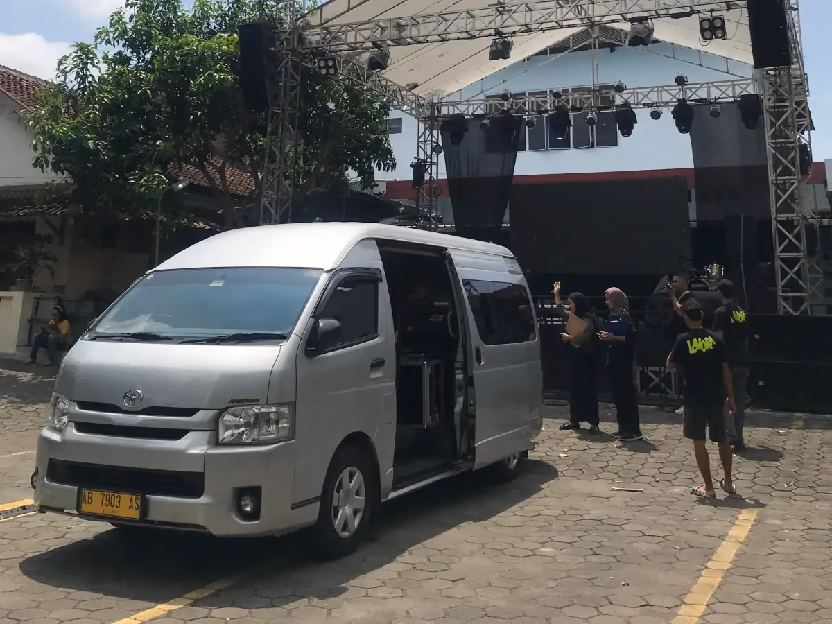 HiAce Commuter silver berdiri di depan panggung acara outdoor dengan crew di sekitar panggung